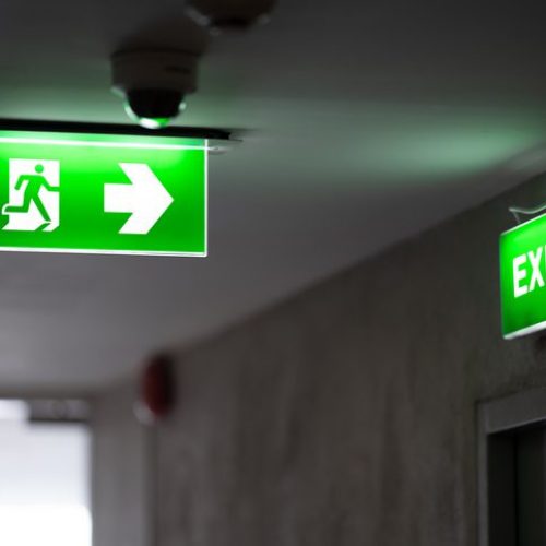 A Green Fire Exit Sign Is Placed On The Ceiling Along The Dimly Lit Corridor And There Is Green Exit Sign On The Exit Do