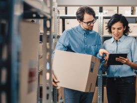 Female Inventory Manager Shows Digital Tablet Information To A Worker Holding Cardboard Box, They Talk And Do Work. In T