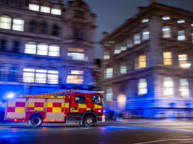 Fast Red Firetruck On Emergency Call In London City Center With Tall Bouldings Around.