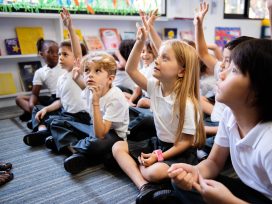 Diverse Group Of Young Students, Boys And Girls, Sit On The Floor In A Classroom Raising Hands.